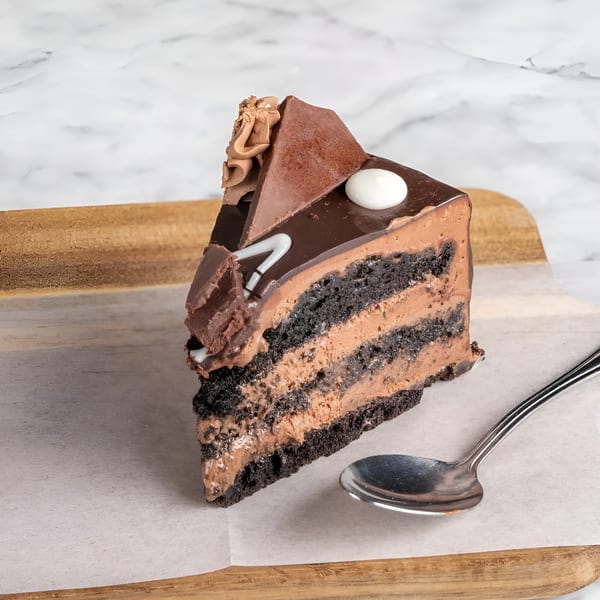 A slice of chocolate cake with chocolate frosting and decorations on a wooden board with a spoon.