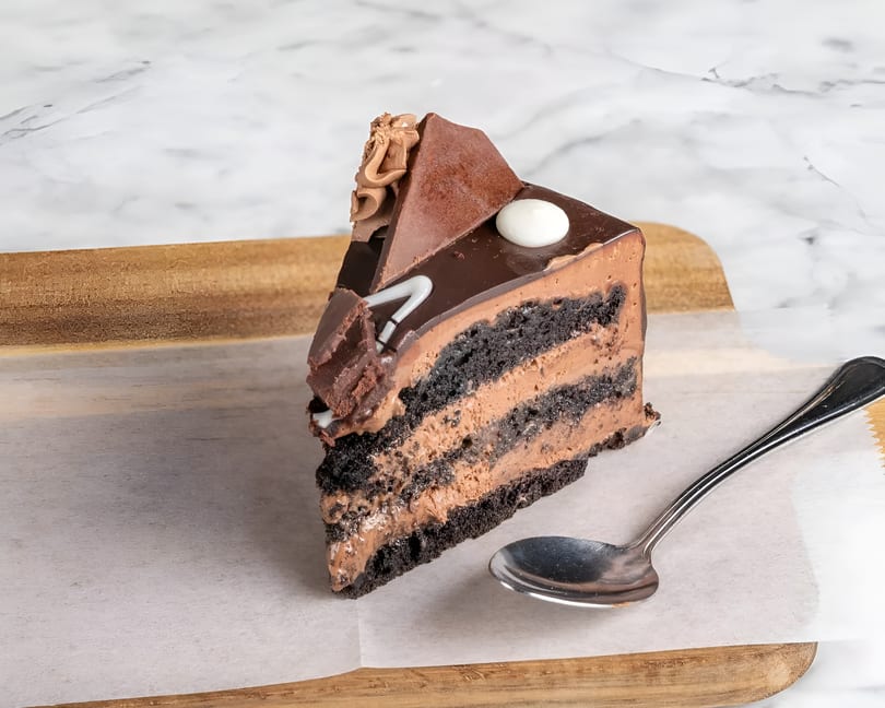 A slice of chocolate cake with chocolate frosting and decorations on a wooden board with a spoon.