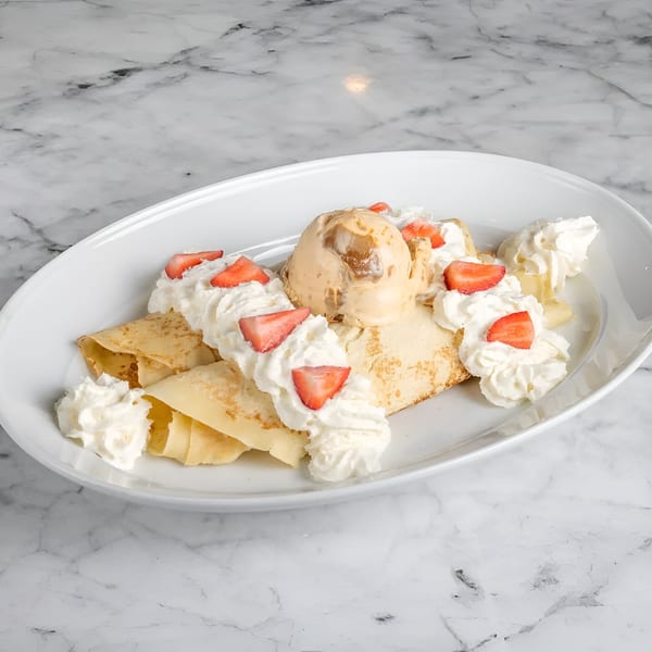 Crepes topped with a scoop of caramel ice cream, whipped cream, and sliced strawberries on a white plate.