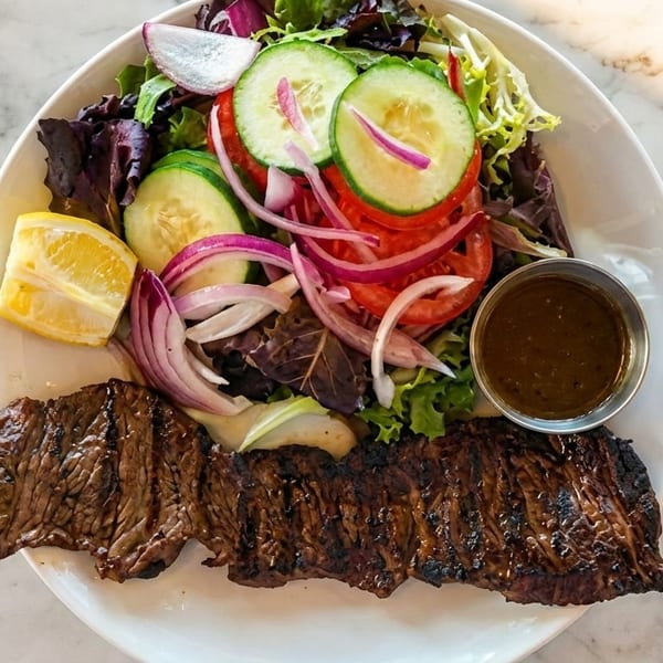 Grilled steak with a side salad of mixed greens, cucumbers, tomatoes, red onions, and a lemon wedge.