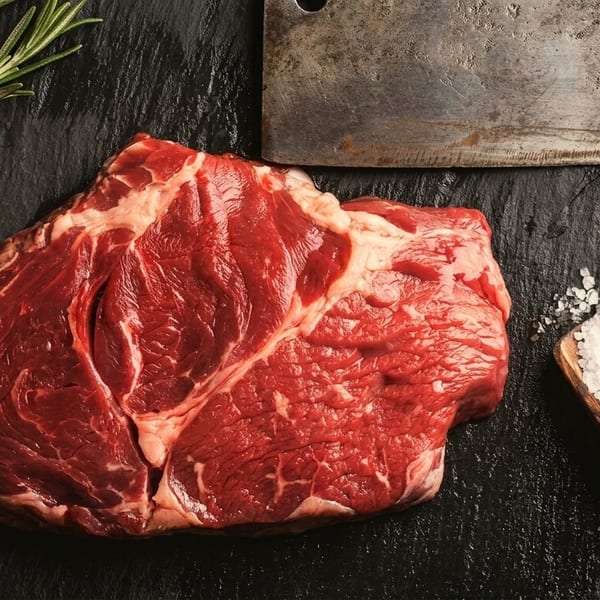 Raw beef steak with rosemary, a meat cleaver, and a wooden scoop of salt on a dark surface.